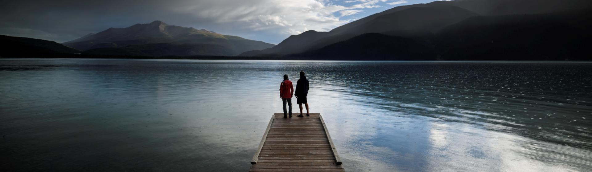 Two people standing on a dock looking towards a lake with low mountains in the background
